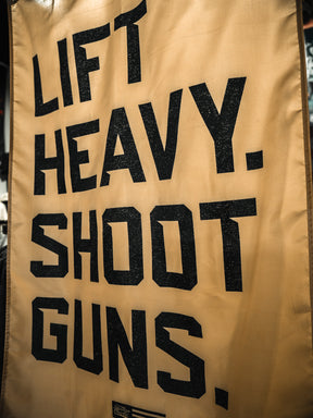 The Lift Heavy. Shoot Guns. Flag by Tango Charlie Apparel features bold black lettering on tan fabric, showcasing your passion for strength training and shooting sports in a striking, eye-catching design.