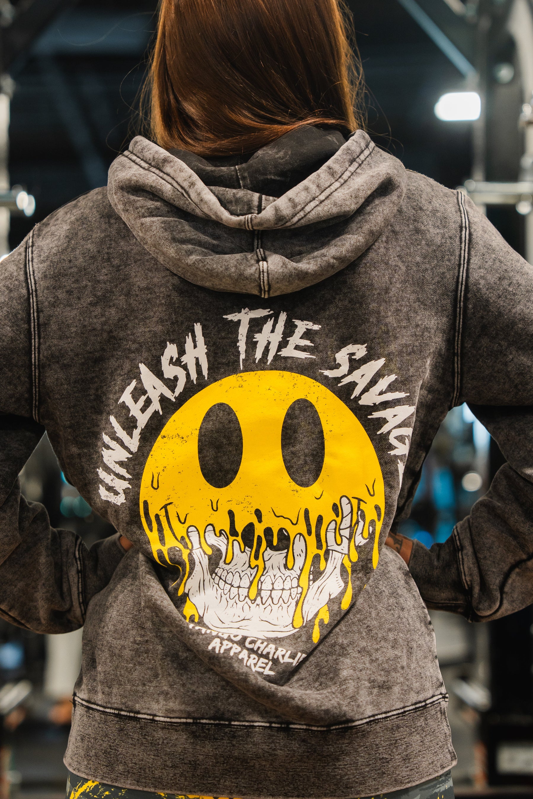A woman faces away in a gray Unleash The Savage Premium Hoodie by Tango Charlie Apparel, featuring a melting smiley over a skull with “UNLEASH THE SAVAGE” above and “TANGO CHARLIE APPAREL” below—embracing your authentic self.
