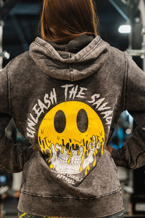 A woman faces away in a gray Unleash The Savage Premium Hoodie by Tango Charlie Apparel, featuring a melting smiley over a skull with “UNLEASH THE SAVAGE” above and “TANGO CHARLIE APPAREL” below—embracing your authentic self.