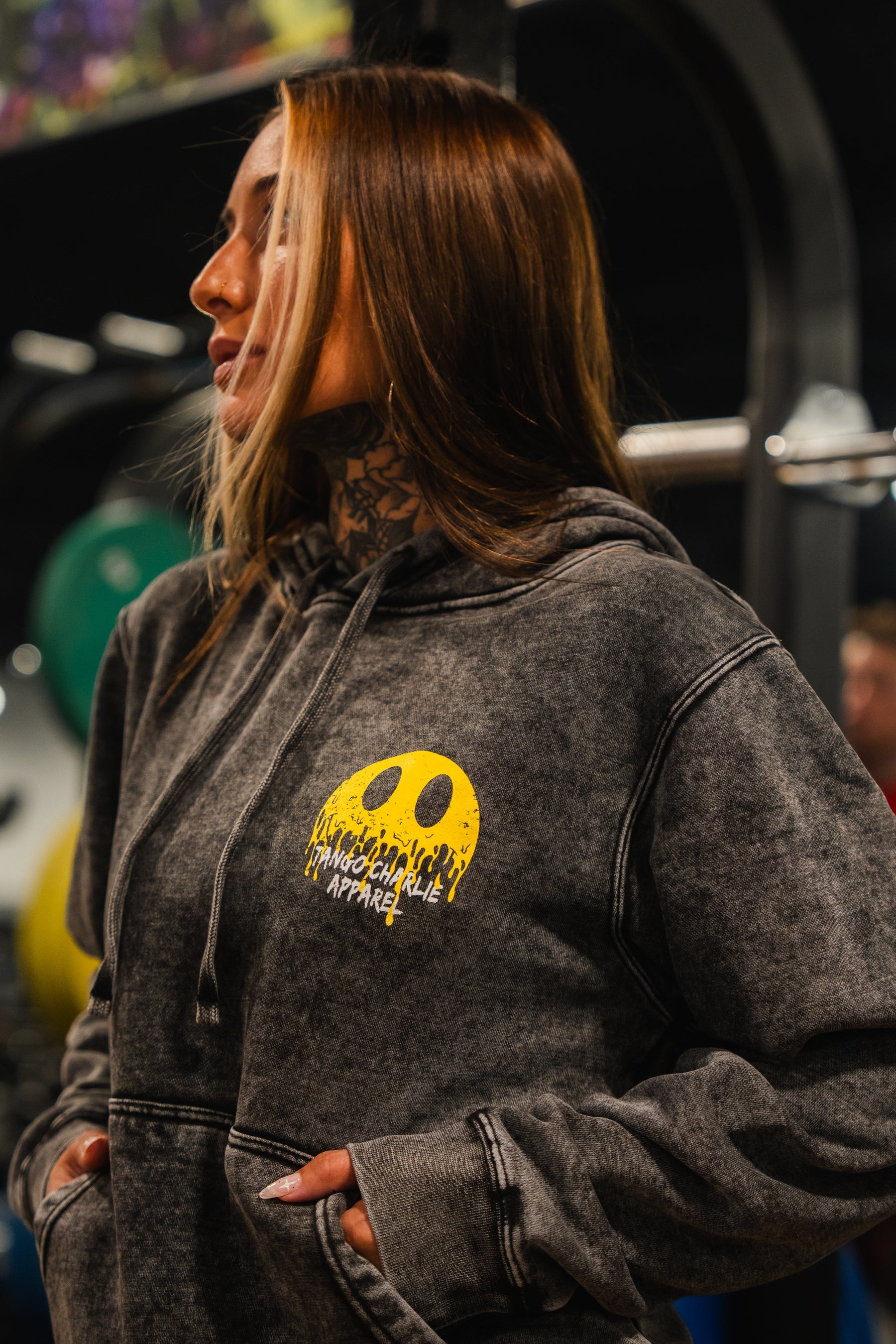 A woman with long hair and neck tattoos wears the Unleash The Savage Premium Hoodie by Tango Charlie Apparel, featuring a melting smile graphic and text, standing indoors with gym equipment in the background.