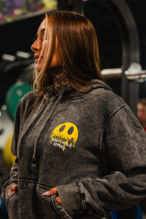 A woman with long hair and neck tattoos wears the Unleash The Savage Premium Hoodie by Tango Charlie Apparel, featuring a melting smile graphic and text, standing indoors with gym equipment in the background.