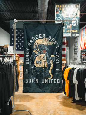A Born United Private Label Honey Badgerin' Flag hangs in a clothing store, featuring a tactical badger and “Honey Badger Don’t Care, Born United.” Clothing racks and American flags fill the bold flag’s background.