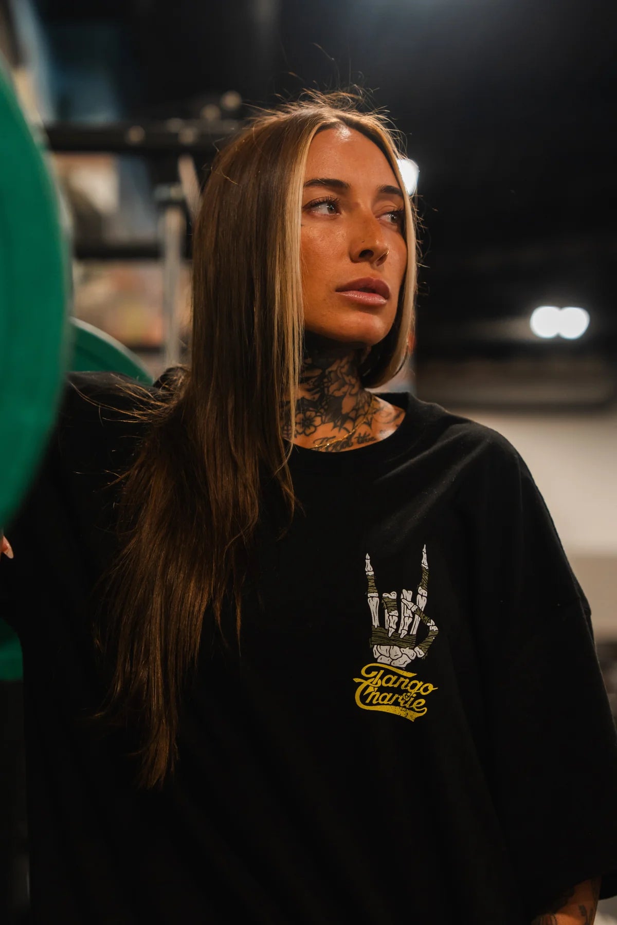 A woman with long brown hair and a blonde streak stands indoors in the Gain Gang Premium Oversized Tee by Tango Charlie Apparel, featuring a graphic hand sign and Charco Charlie text. She has neck tattoos and gazes thoughtfully to the side.