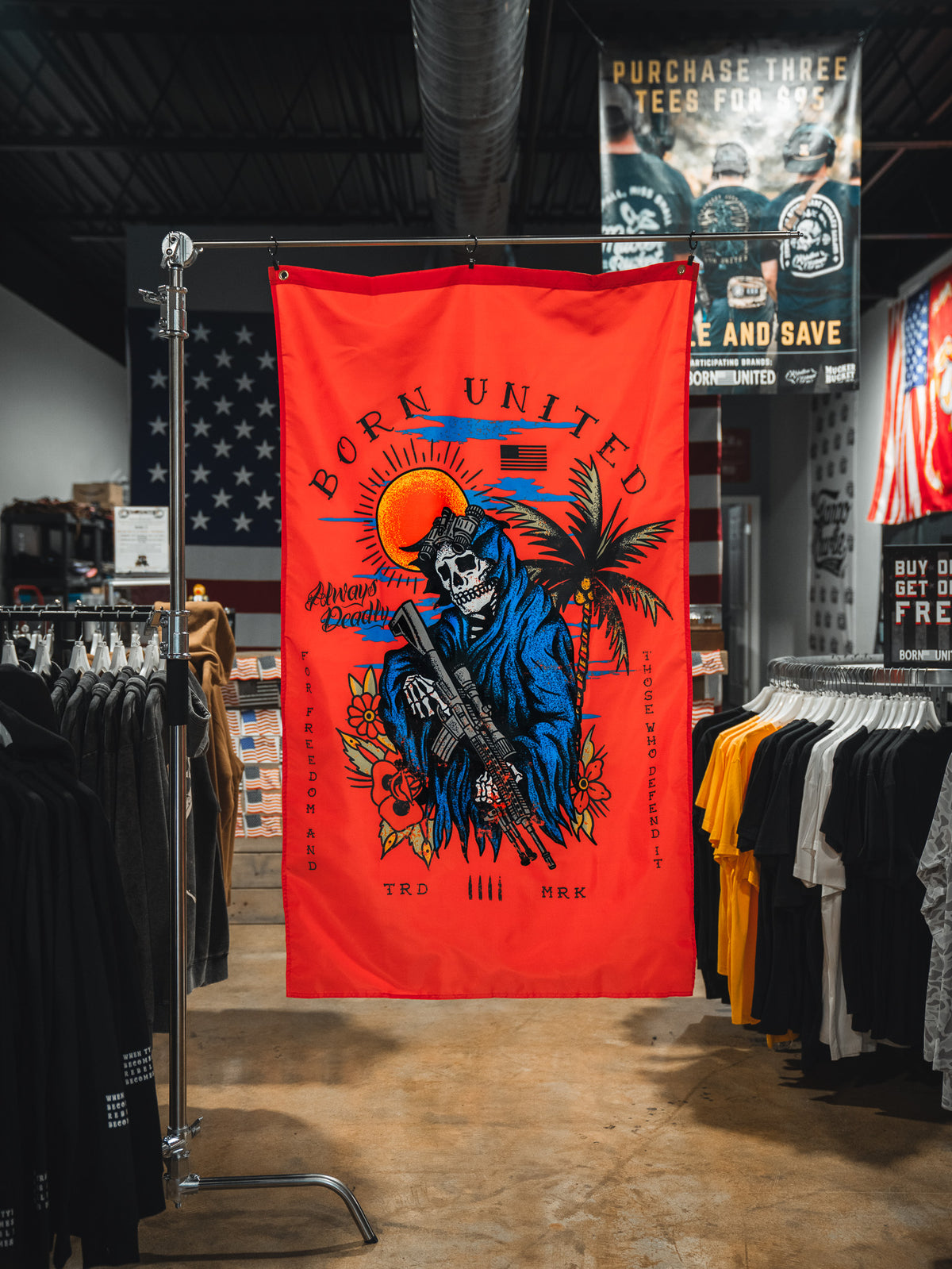 A red Always Deadly Flag by Born United Private Label, featuring a colorful skeleton in a blue cloak with a rifle, palm trees, and a setting sun, hangs in the clothing store. The banner reads "Born United" and "Anatomy of Peaceful.
