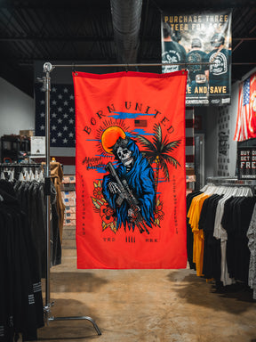 A red Always Deadly Flag by Born United Private Label, featuring a colorful skeleton in a blue cloak with a rifle, palm trees, and a setting sun, hangs in the clothing store. The banner reads "Born United" and "Anatomy of Peaceful.
