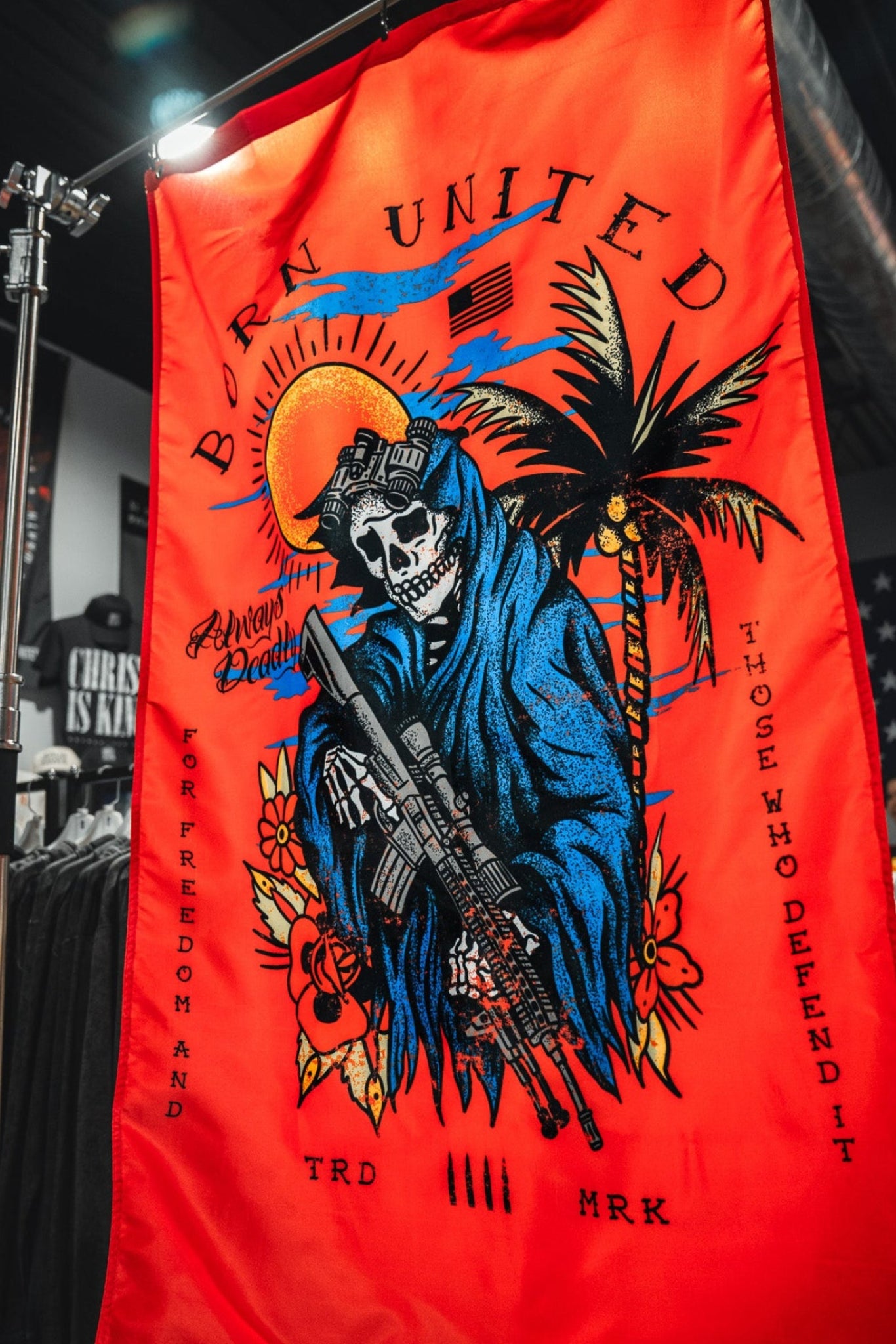 The Always Deadly Flag by Born United Private Label features a red background, a skeleton in a blue hooded robe with a rifle, palm trees, sun, and the text "Born United, For Freedom and Those Who Defend It" plus other small phrases.