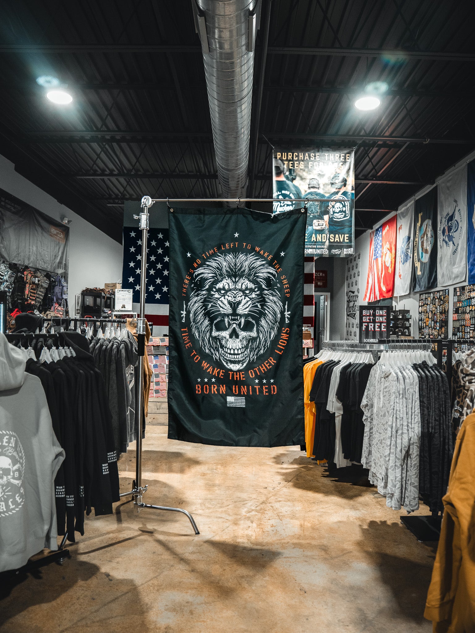 A clothing store showcases the Born United Private Label Waking Lions Flag, featuring a roaring lion and the phrase “It takes one left to wake the others. Born United,” displayed among shirts and hoodies under bright lights.
