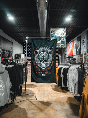 A clothing store showcases the Born United Private Label Waking Lions Flag, featuring a roaring lion and the phrase “It takes one left to wake the others. Born United,” displayed among shirts and hoodies under bright lights.