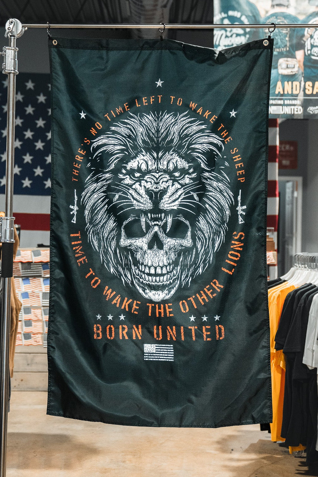 The Born United Private Label Waking Lions Flag features a black background with a roaring lion and skull, surrounded by the text: "There’s no time left to wake the sheep. Time to wake the other lions." T-shirts and American flags appear behind it.