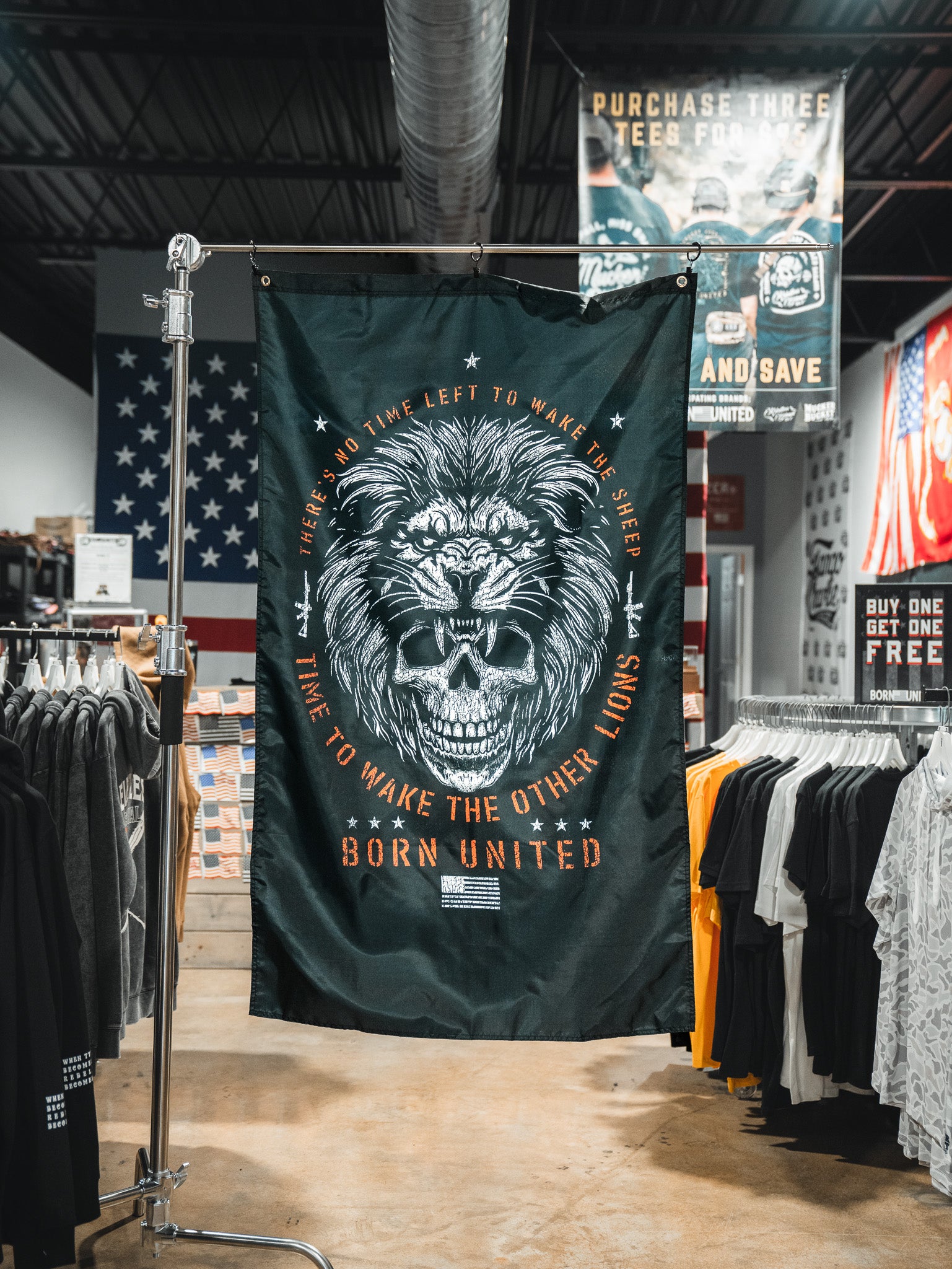 The Born United Private Label Waking Lions Flag features a black background with a roaring lion and skull, surrounded by the text: "There’s no time left to wake the sheep. Time to wake the other lions." T-shirts and American flags appear behind it.
