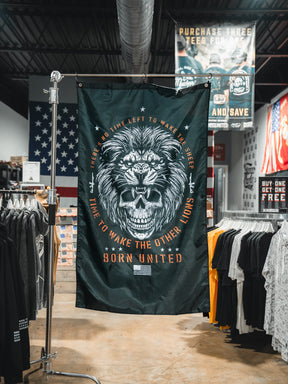 The Born United Private Label Waking Lions Flag features a black background with a roaring lion and skull, surrounded by the text: "There’s no time left to wake the sheep. Time to wake the other lions." T-shirts and American flags appear behind it.