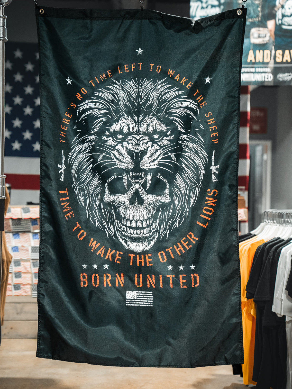 The Waking Lions Flag by Born United Private Label, crafted from durable polyester, showcases a roaring lion over a skull with the text: “There’s no time left to wake the sheep. Time to wake the other lions. Born United.” Displayed in a store setting.