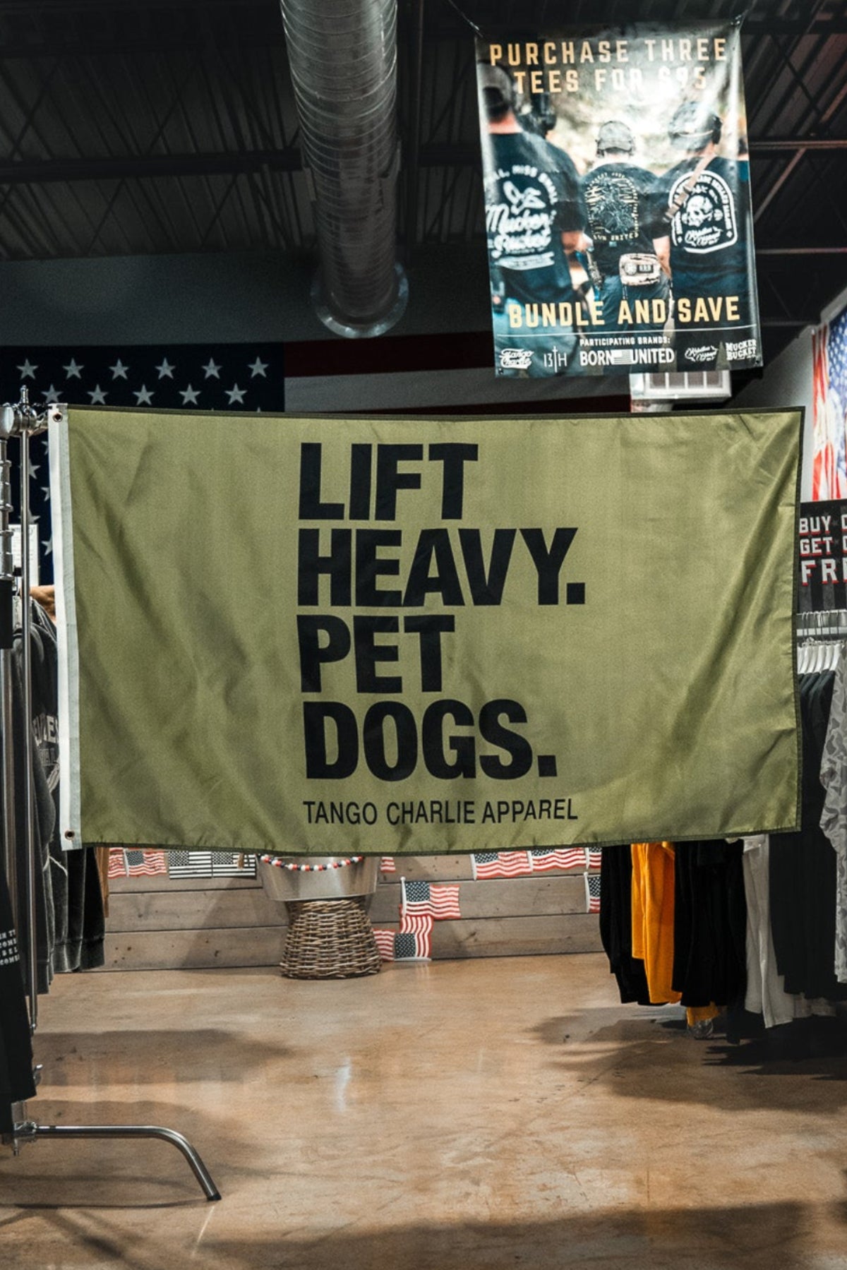 A large olive green banner hanging in the center of a clothing store aisle, printed with the text 'LIFT HEAVY. PET DOGS.
