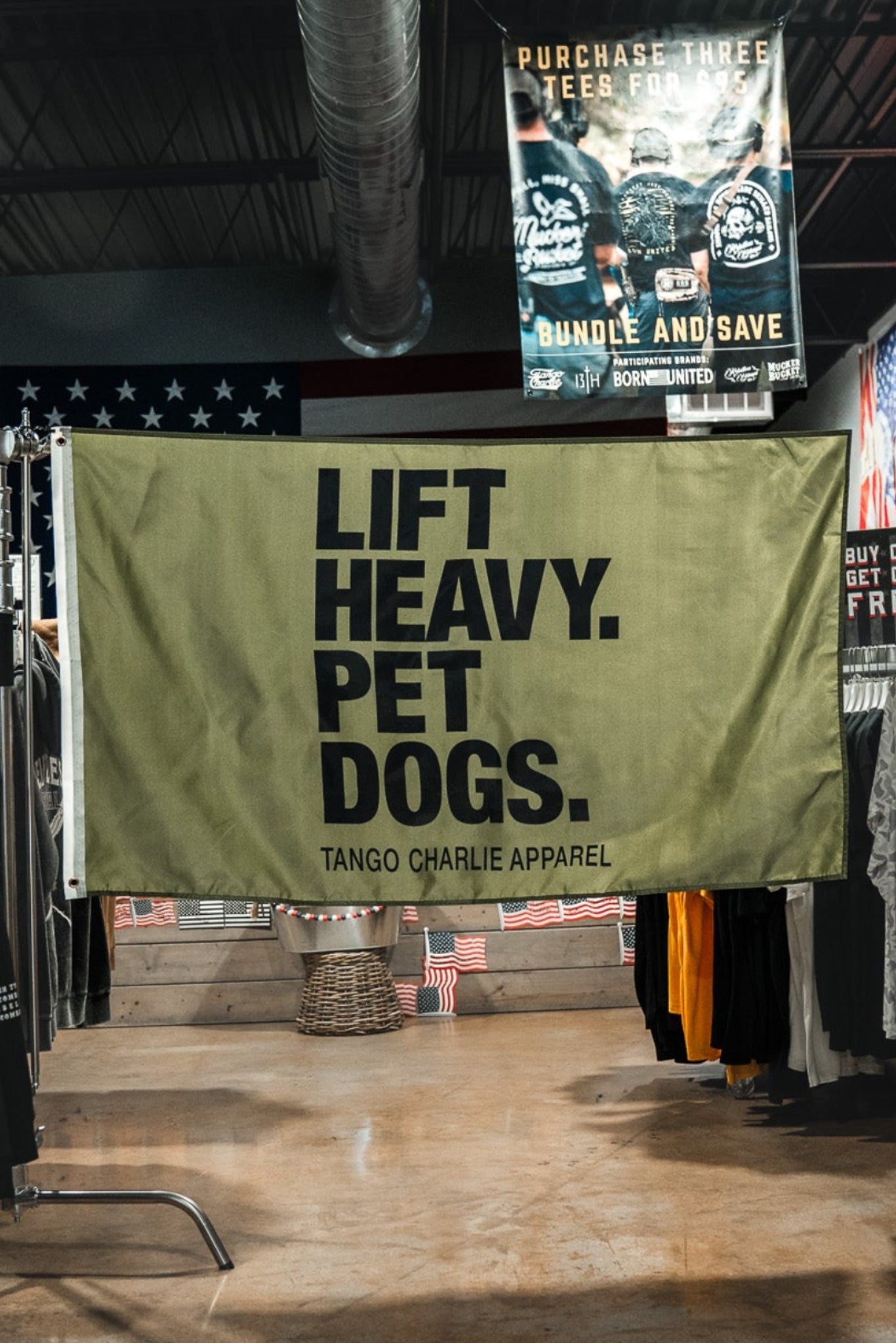 A large olive green banner hanging in the center of a clothing store aisle, printed with the text 'LIFT HEAVY. PET DOGS.