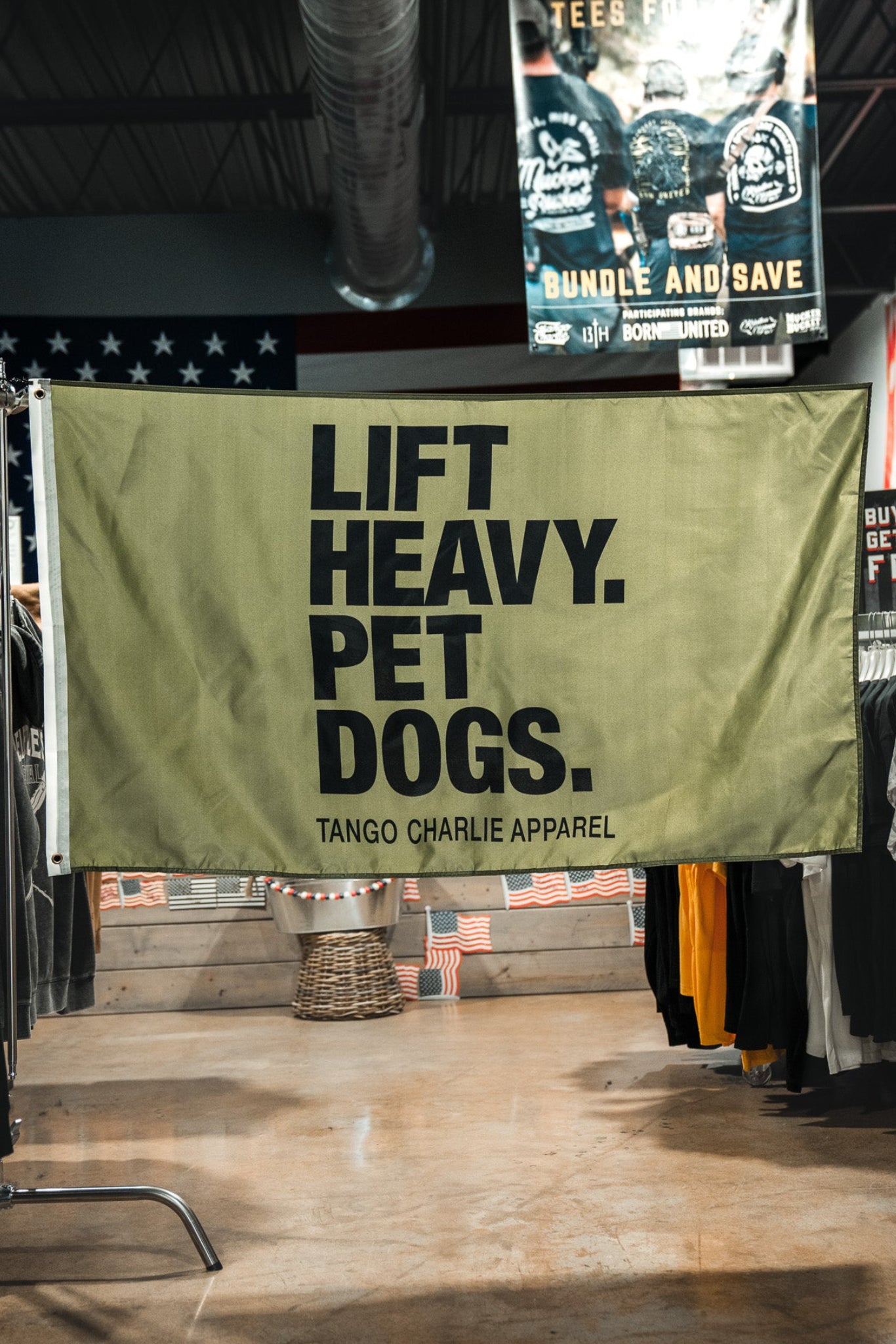 A large olive green fabric banner hanging in a retail store, printed with the bold black text 'LIFT HEAVY. PET DOGS.