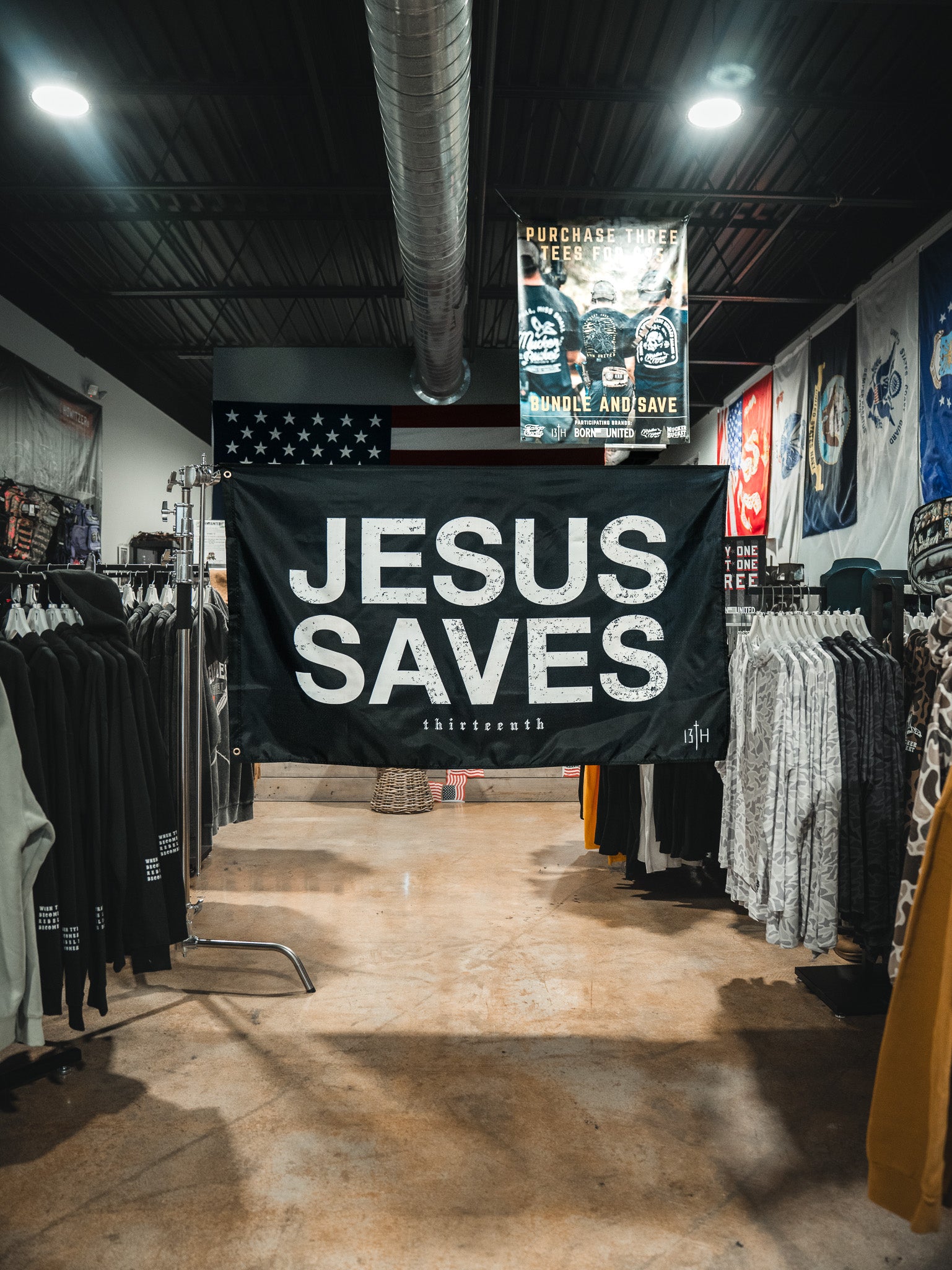 A large 13th Jesus Saves Flag - Black with white lettering hangs in the center of a clothing store, surrounded by racks of clothes and colorful wall banners.