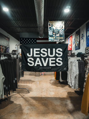 A large 13th Jesus Saves Flag - Black with white lettering hangs in the center of a clothing store, surrounded by racks of clothes and colorful wall banners.