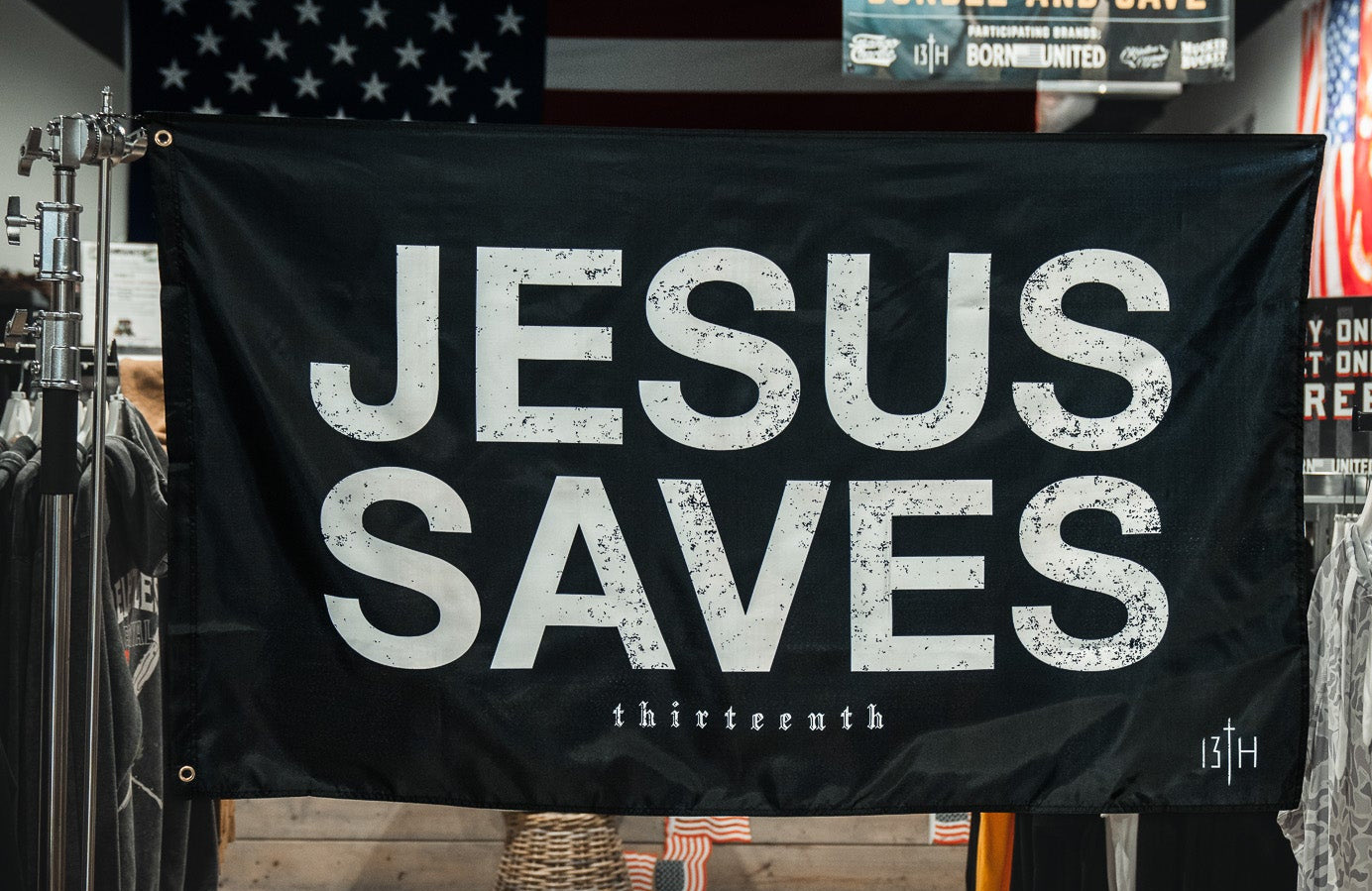 A 13th Jesus Saves Flag - Black with bold white text hangs in a clothing store, surrounded by shirt racks. An American flag and a promotional sign are also visible in the background.