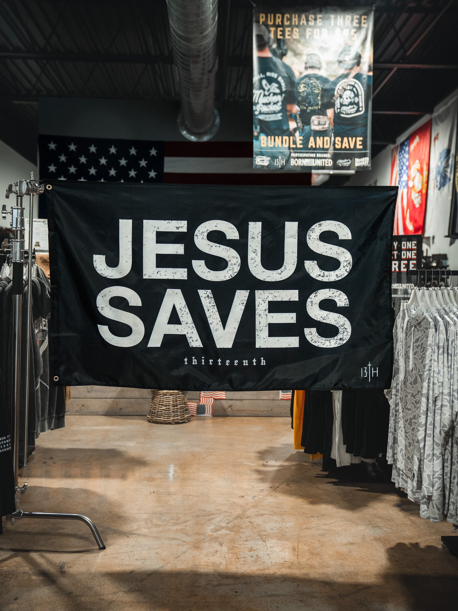 A 13th Jesus Saves Flag - Black with bold white text hangs in a clothing store, surrounded by shirt racks. An American flag and a promotional sign are also visible in the background.