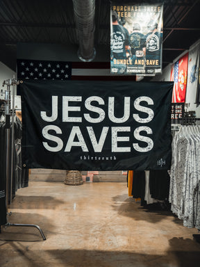 A 13th Jesus Saves Flag - Black with bold white text hangs in a clothing store, surrounded by shirt racks. An American flag and a promotional sign are also visible in the background.