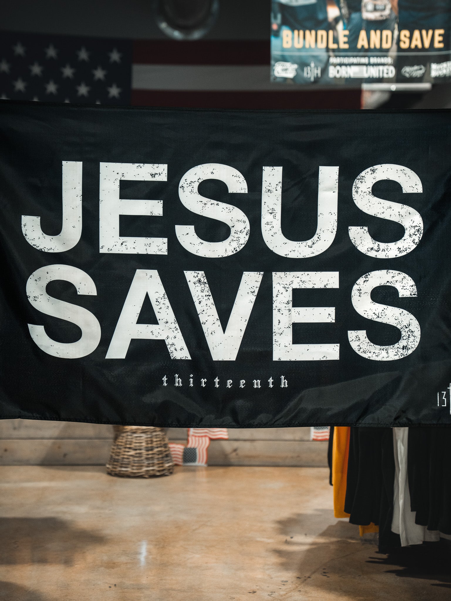 The Jesus Saves Flag - Black by 13th features bold distressed white JESUS SAVES text with "thirteenth" below. The flag is displayed indoors among store items, with a blurred American flag seen in the background.