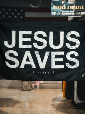 The Jesus Saves Flag - Black by 13th features bold distressed white JESUS SAVES text with "thirteenth" below. The flag is displayed indoors among store items, with a blurred American flag seen in the background.