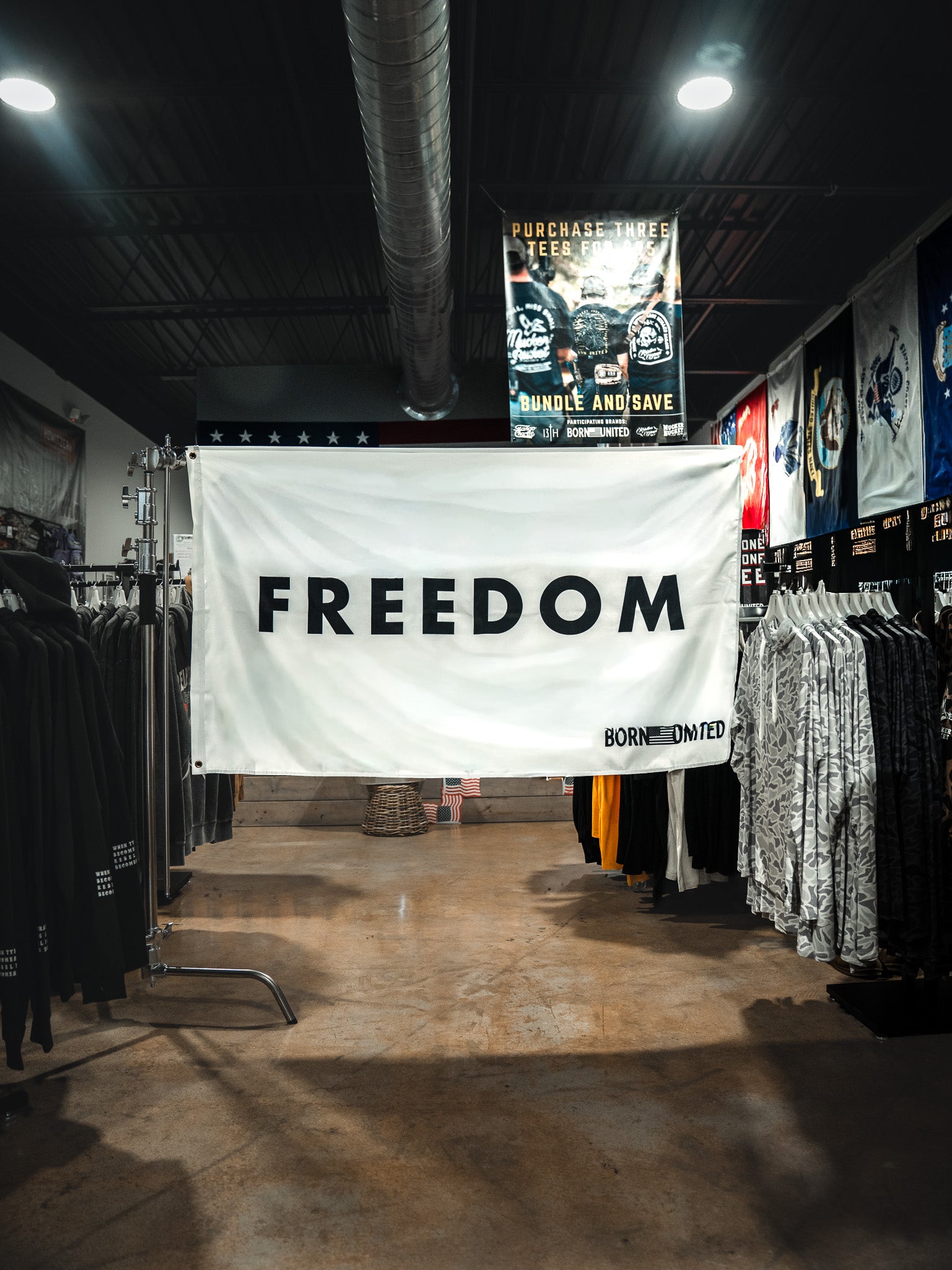 A large white Freedom Flag by Born United Private Label with bold black "FREEDOM" lettering hangs between clothing racks in a store, reflecting patriotic values. Various shirts and posters can be seen in the background.