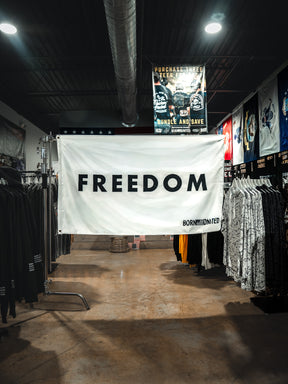 A large white Freedom Flag by Born United Private Label with bold black "FREEDOM" lettering hangs between clothing racks in a store, reflecting patriotic values. Various shirts and posters can be seen in the background.