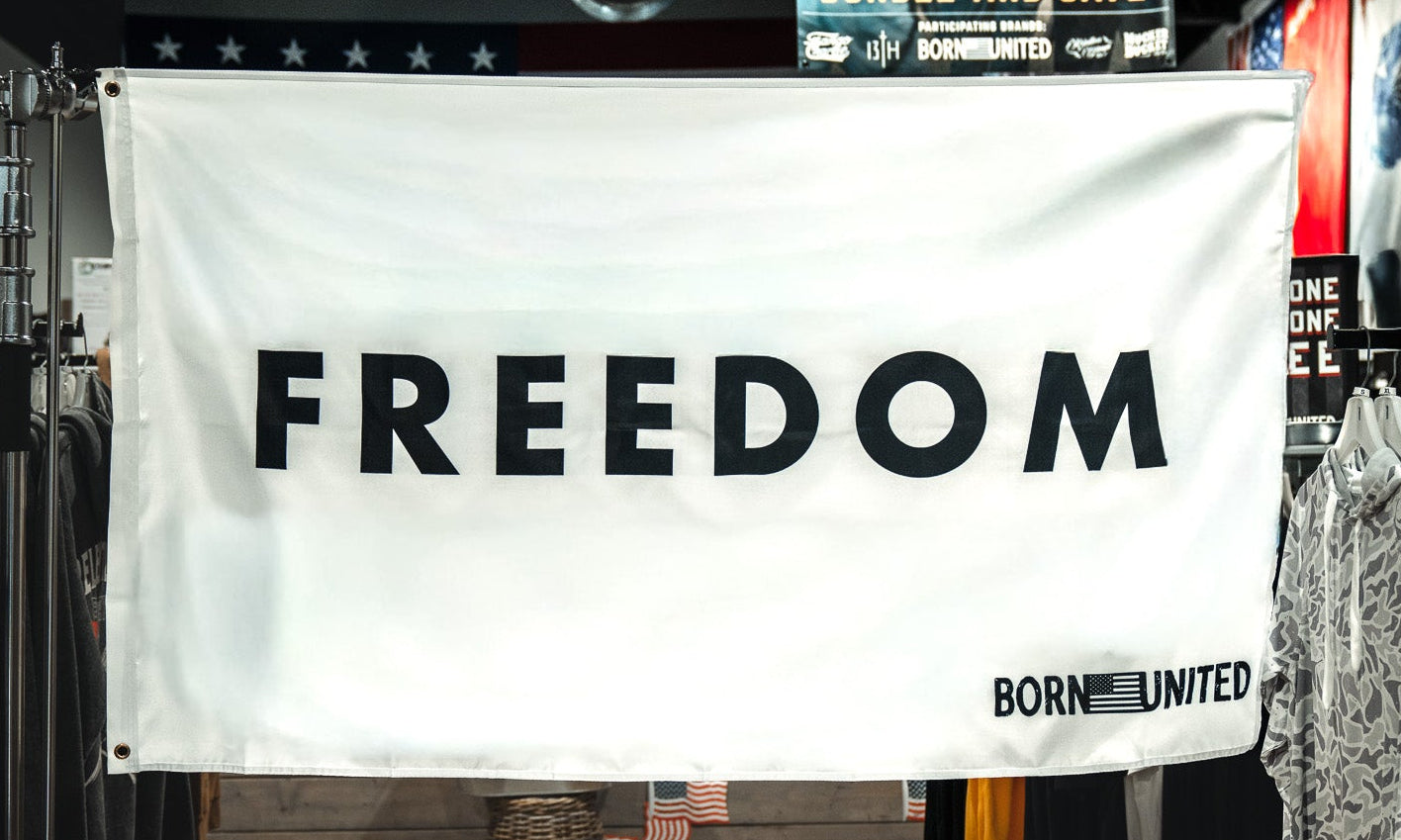 In a clothing store, a large white Freedom Flag by Born United Private Label hangs boldly with "FREEDOM" in black letters and "BORN UNITED" in the corner, surrounded by racks of shirts and evoking patriotic values.