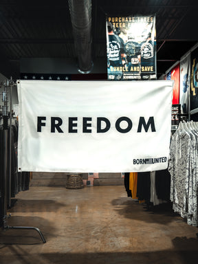 In a clothing store, a large white Freedom Flag by Born United Private Label hangs boldly with "FREEDOM" in black letters and "BORN UNITED" in the corner, surrounded by racks of shirts and evoking patriotic values.
