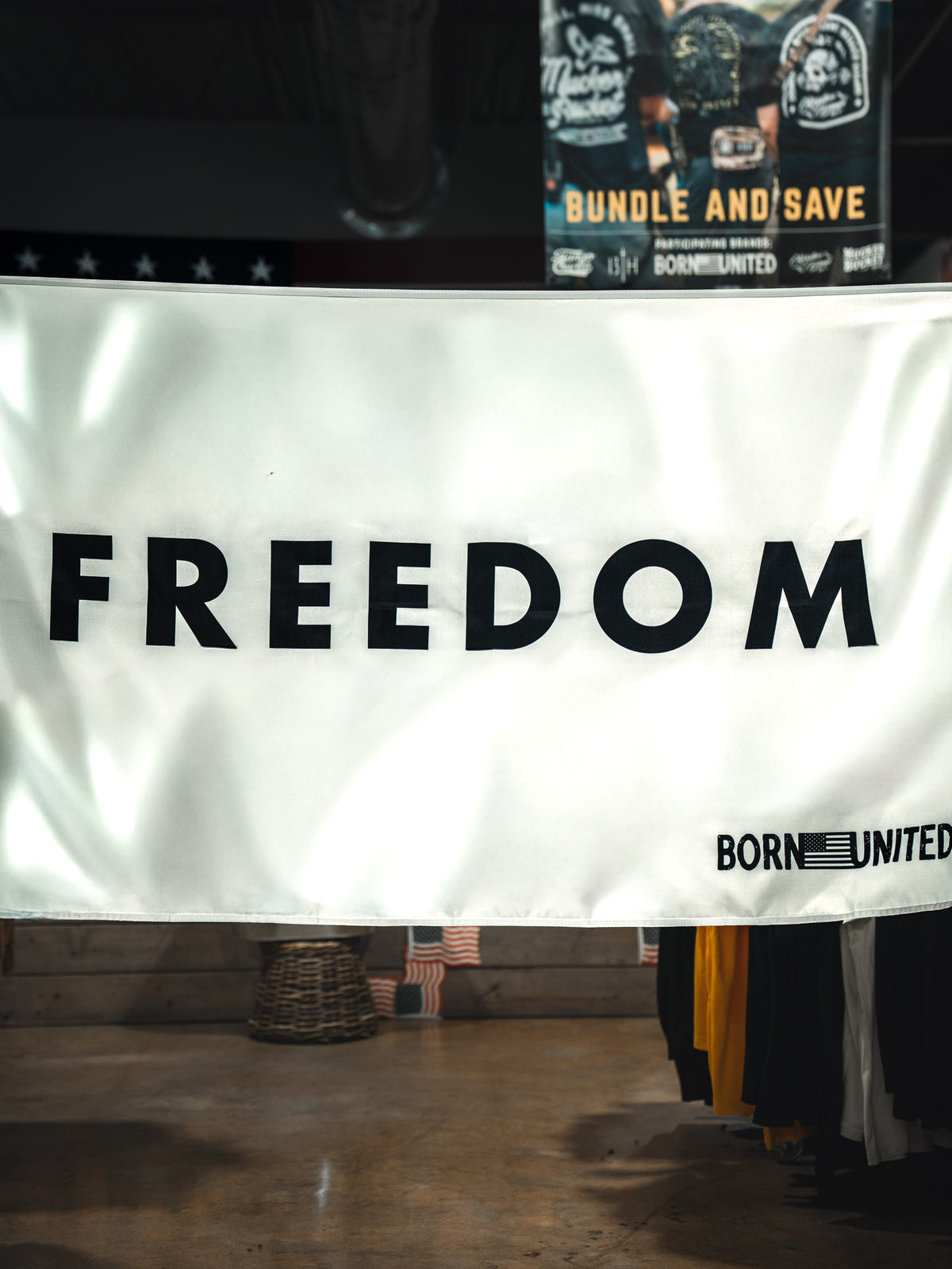 A white Freedom Flag by Born United Private Label hangs indoors with bold black "FREEDOM" and an American flag corner featuring "BORN UNITED," embodying patriotism. Clothing racks and a store sign appear in the background.