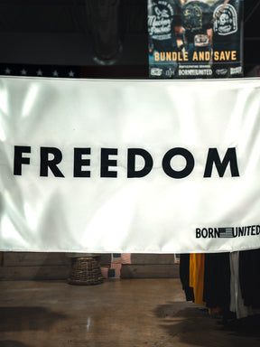 A white Freedom Flag by Born United Private Label hangs indoors with bold black "FREEDOM" and an American flag corner featuring "BORN UNITED," embodying patriotism. Clothing racks and a store sign appear in the background.