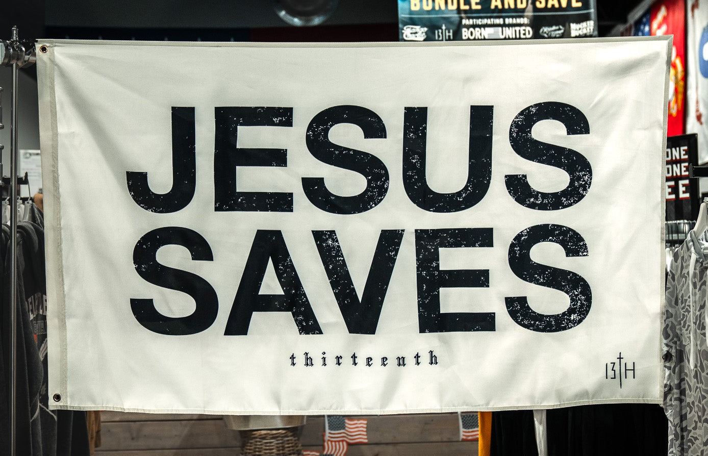 An ivory "Jesus Saves Flag" by 13th with bold black text hangs in a clothing store, surrounded by shirts and racks. A promotional poster is visible in the background.