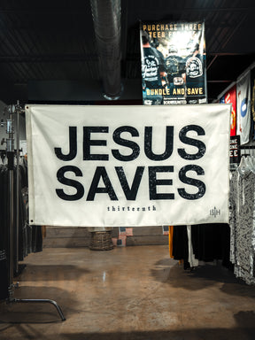 An ivory "Jesus Saves Flag" by 13th with bold black text hangs in a clothing store, surrounded by shirts and racks. A promotional poster is visible in the background.