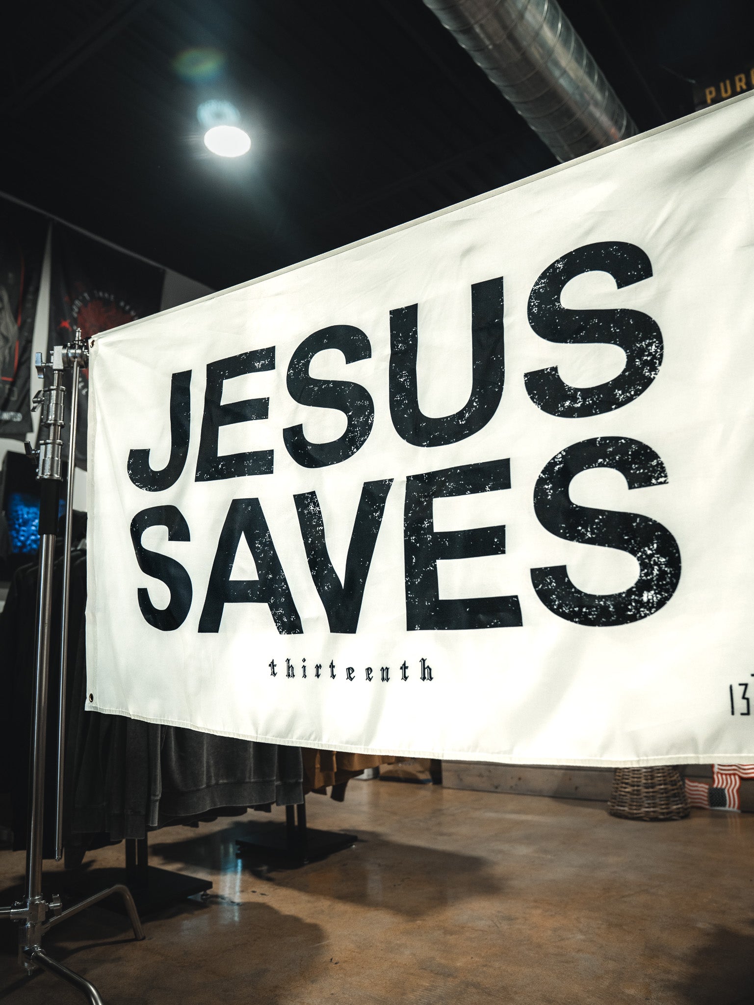 The Jesus Saves Flag - Ivory by 13th features bold, distressed black letters and "thirteenth" in small text below. Display it on a metal stand indoors for faith-inspired home decor or outdoors as a unique Christian statement flag.