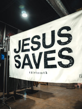 The Jesus Saves Flag - Ivory by 13th features bold, distressed black letters and "thirteenth" in small text below. Display it on a metal stand indoors for faith-inspired home decor or outdoors as a unique Christian statement flag.