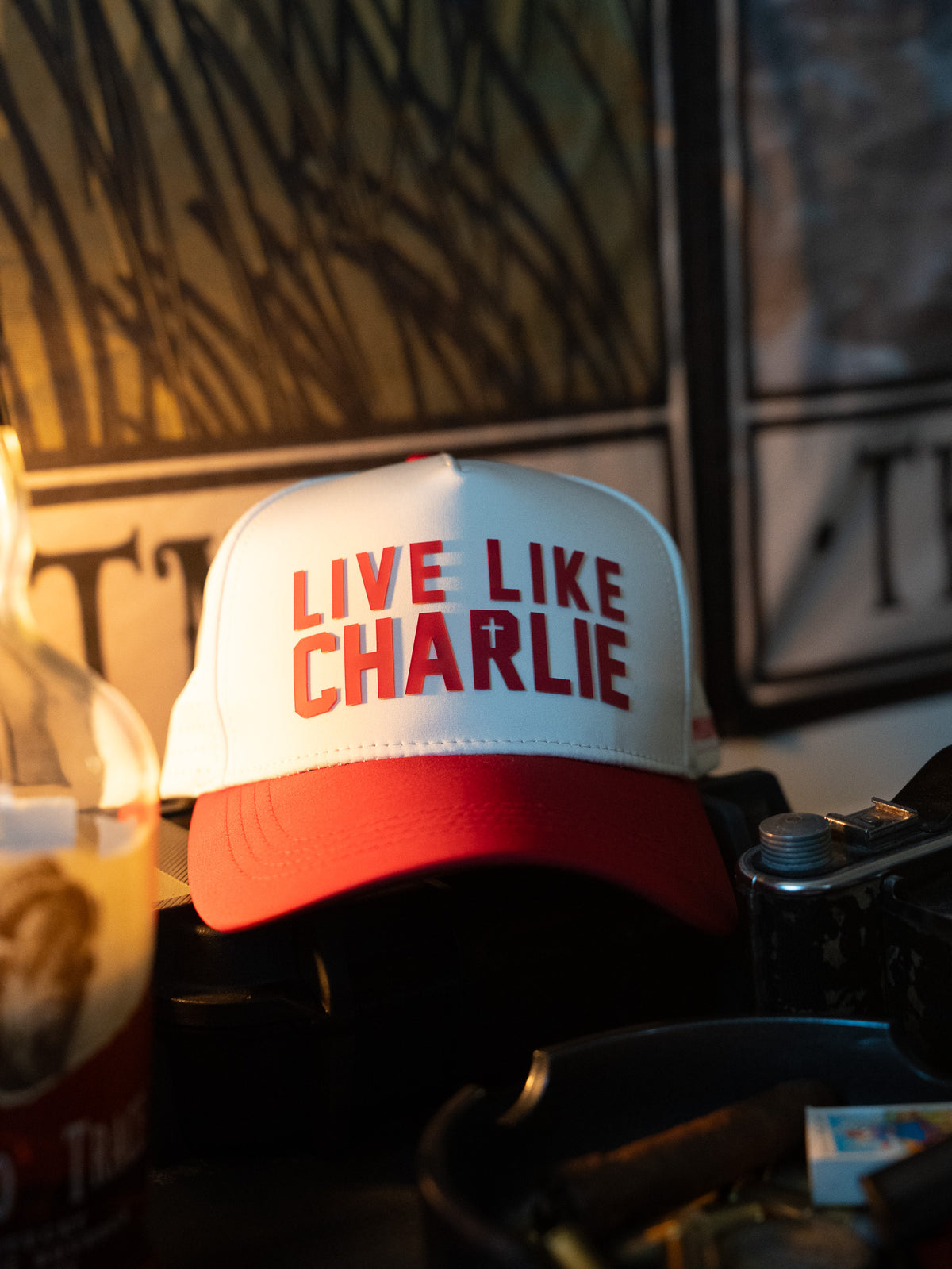 The Born United Private Label Live Like Charlie Ivory Snapback, featuring "LIVE LIKE CHARLIE" and a small cross on the front, rests on a dark surface amid various objects in dim lighting.