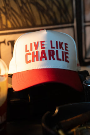 The Born United Private Label Live Like Charlie Ivory Snapback, featuring "LIVE LIKE CHARLIE" and a small cross on the front, rests on a dark surface amid various objects in dim lighting.