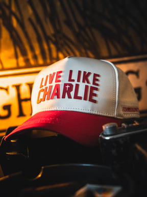 The Born United Private Label Live Like Charlie Ivory Snapback, featuring bold red lettering, is stylishly displayed against a warm, blurred background for a fashionable and comfortable look.