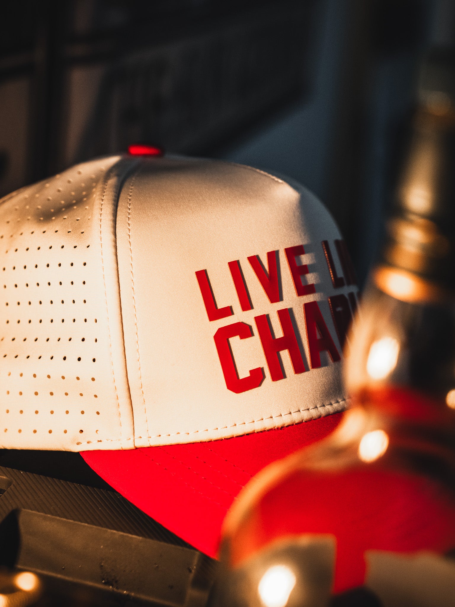 The Born United Private Label Live Like Charlie Ivory Snapback is a white and red 5-panel hat with bold red "LIVE LIKE CHARLIE" text, shown on a warmly lit surface. This breathable snapback adds a stylish touch.