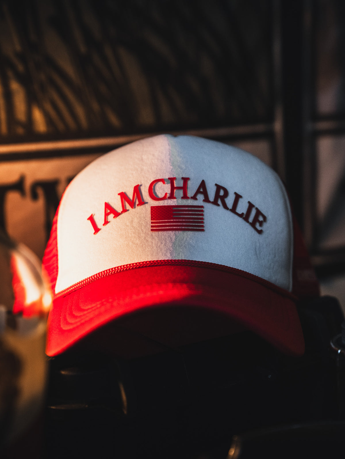The Born United Private Label I Am Charlie Red Trucker Snapback features a red and white 5-panel curved brim, "I AM CHARLIE" text, and a stylized American flag on the front, shown in focus resting indoors against a dark background.