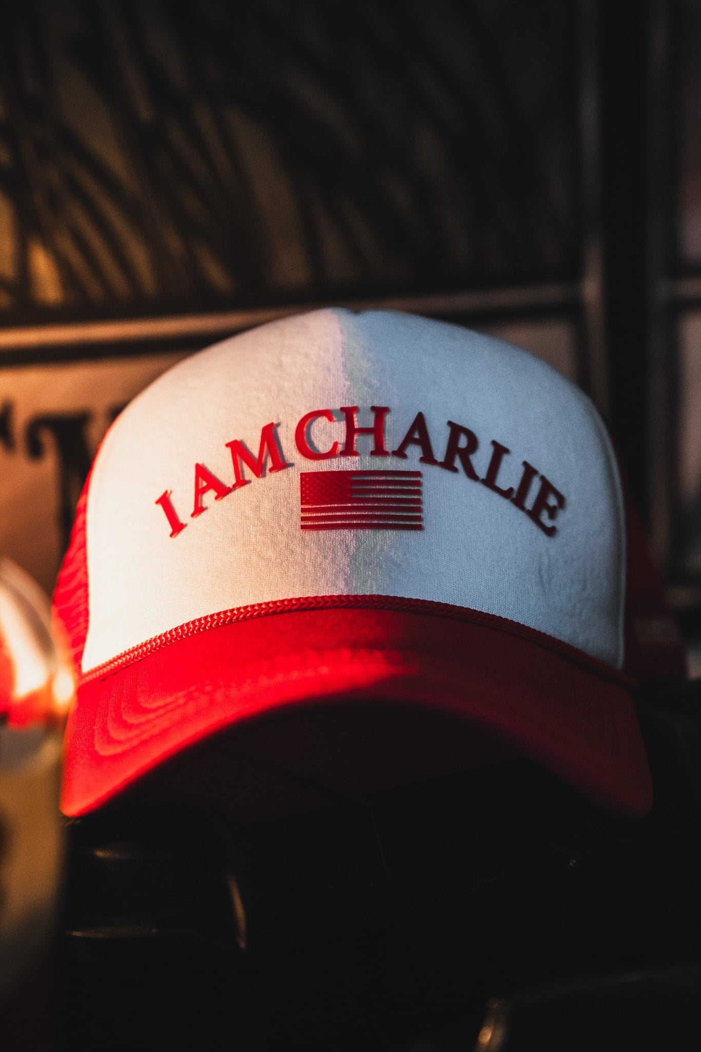 The Born United Private Label I Am Charlie Red Trucker Snapback features a red and white 5-panel curved brim, "I AM CHARLIE" text, and a stylized American flag on the front, shown in focus resting indoors against a dark background.