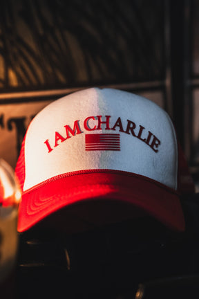 The Born United Private Label I Am Charlie Red Trucker Snapback features a red and white 5-panel curved brim, "I AM CHARLIE" text, and a stylized American flag on the front, shown in focus resting indoors against a dark background.