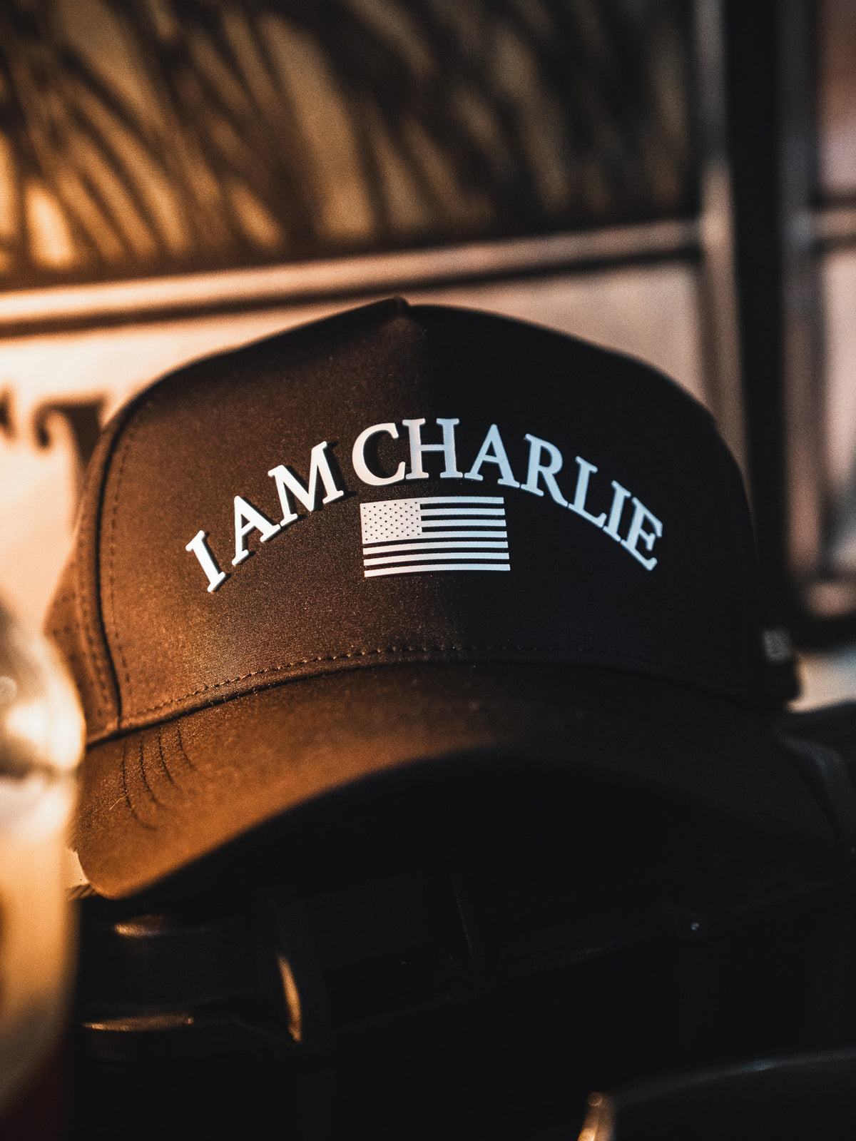 The I Am Charlie Black Snapback by Born United Private Label is a handmade, breathable baseball cap featuring "I AM CHARLIE" in white above an American flag graphic. Shown resting on a dark surface with a blurred background.