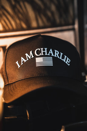 The I Am Charlie Black Snapback by Born United Private Label is a handmade, breathable baseball cap featuring "I AM CHARLIE" in white above an American flag graphic. Shown resting on a dark surface with a blurred background.