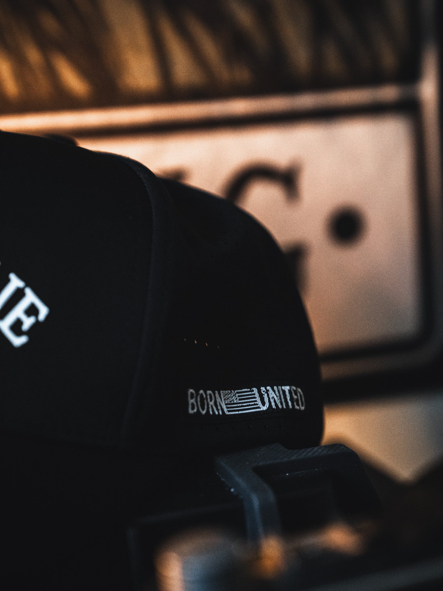 A close-up of the I Am Charlie Black Snapback from Born United Private Label, featuring "BORN UNITED" text and an American flag stitched on the side, set against a blurred background with warm lighting.