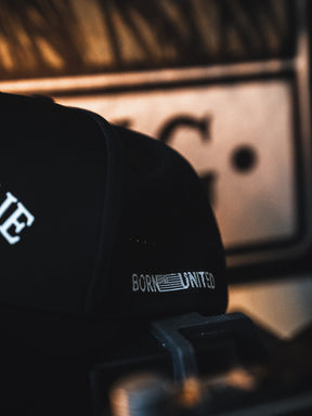 A close-up of the I Am Charlie Black Snapback from Born United Private Label, featuring "BORN UNITED" text and an American flag stitched on the side, set against a blurred background with warm lighting.