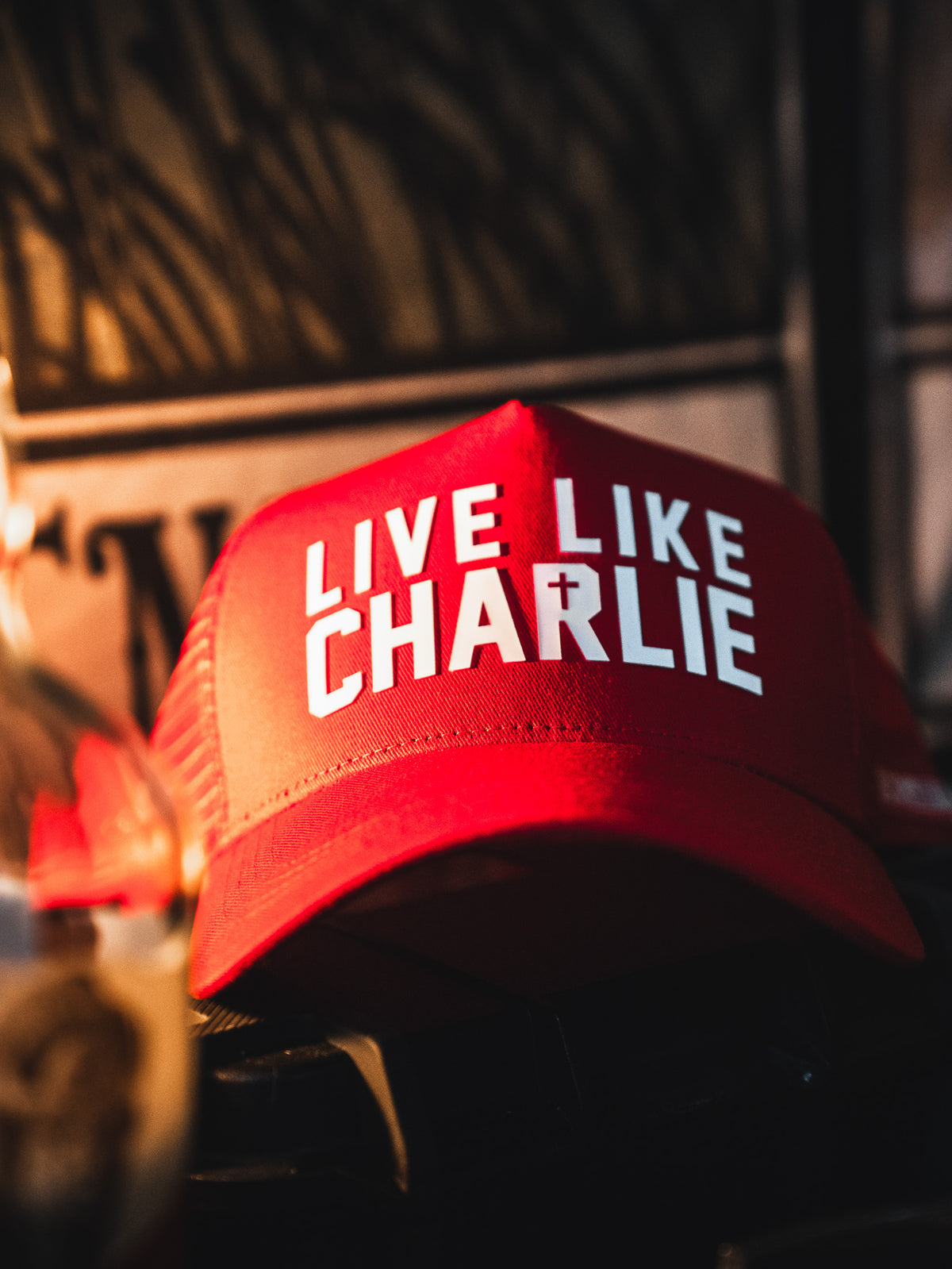 The Born United Private Label "Live Like Charlie Trucker Snapback" is a red 5-panel cap with a curved brim and bold white “LIVE LIKE CHARLIE” lettering on the front, shown indoors with warm lighting and a blurred background.