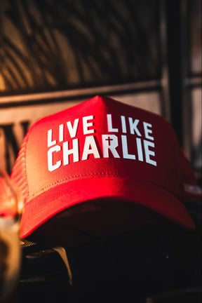 The Born United Private Label "Live Like Charlie Trucker Snapback" is a red 5-panel cap with a curved brim and bold white “LIVE LIKE CHARLIE” lettering on the front, shown indoors with warm lighting and a blurred background.