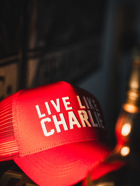 The Born United Private Label "Live Like Charlie Trucker Snapback" is a red mesh 5-panel hat with bold white text, a cross as the I in CHARLIE, and classic snapback design shown under warm light.